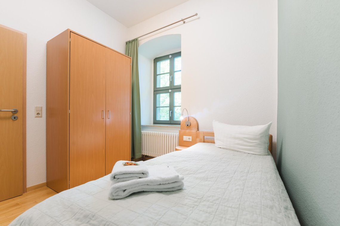 Ein kleines Zimmer mit einem Fenster, einem Bett mit 2 Handtüchern, einem Schrank und einer Tür.