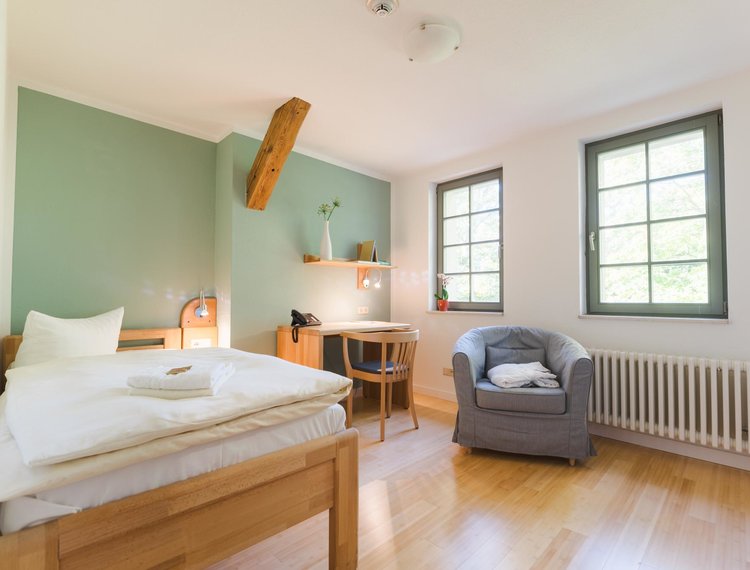 Man sieht ein Hotelzimmer mit links einem Bett mit Handtüchern, in der Mitte einem Schreibtisch mit einem Stuhl und einem Bücherregal darüber und rechts daneben einen grauen Sessel vor zwei Fenstern. Die Mittlere Wand ist grün. An der Decke sieht man einen Feuermelder und eine Lampe.