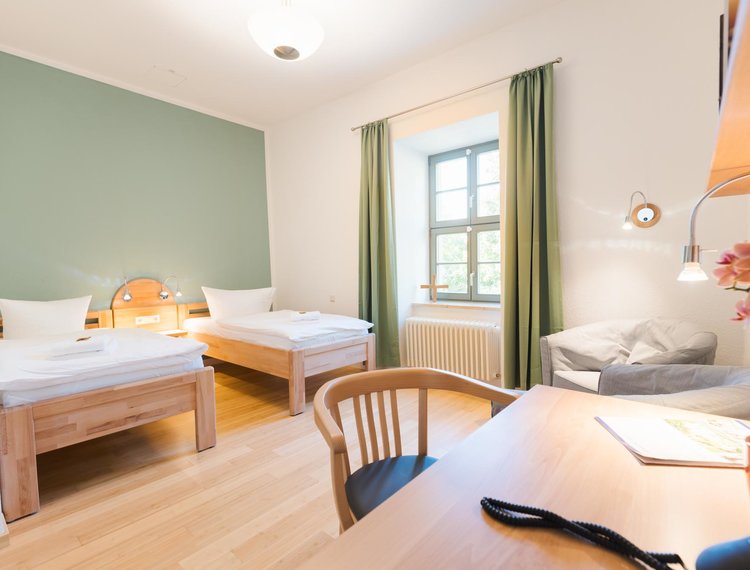 Ein helles weißes Zimmer mit einem Fenster mit grünen Vorhängen. Im Vordergrund steht ein Schreibtisch mit einem Stuhl. Auf dem Schreibtisch steht ein Telefon und eine Blume. Über dem Schreibtisch ist ein Bücherregal unter dem eine Lampe hängt. Hinter dem Schreibtisch stehen zwei graue Sessel. An einer grünen Wand stehen zwei Einzelbetten mit zwei Lampen dazwischen.