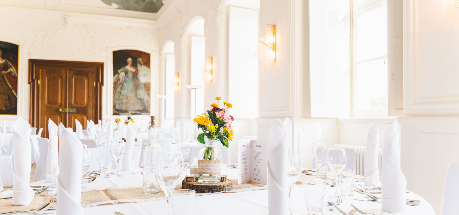 Der große Kaisersaal ist feierlich geschmückt. Die runden Tische sind mit weißen Tischdecken gedeckt und die Stühle in weißen Stoffen gehüllt. Die Tische sind gedeckt mit Besteck, weißen Servietten, einer Blumenvase und der Buffetkarte.