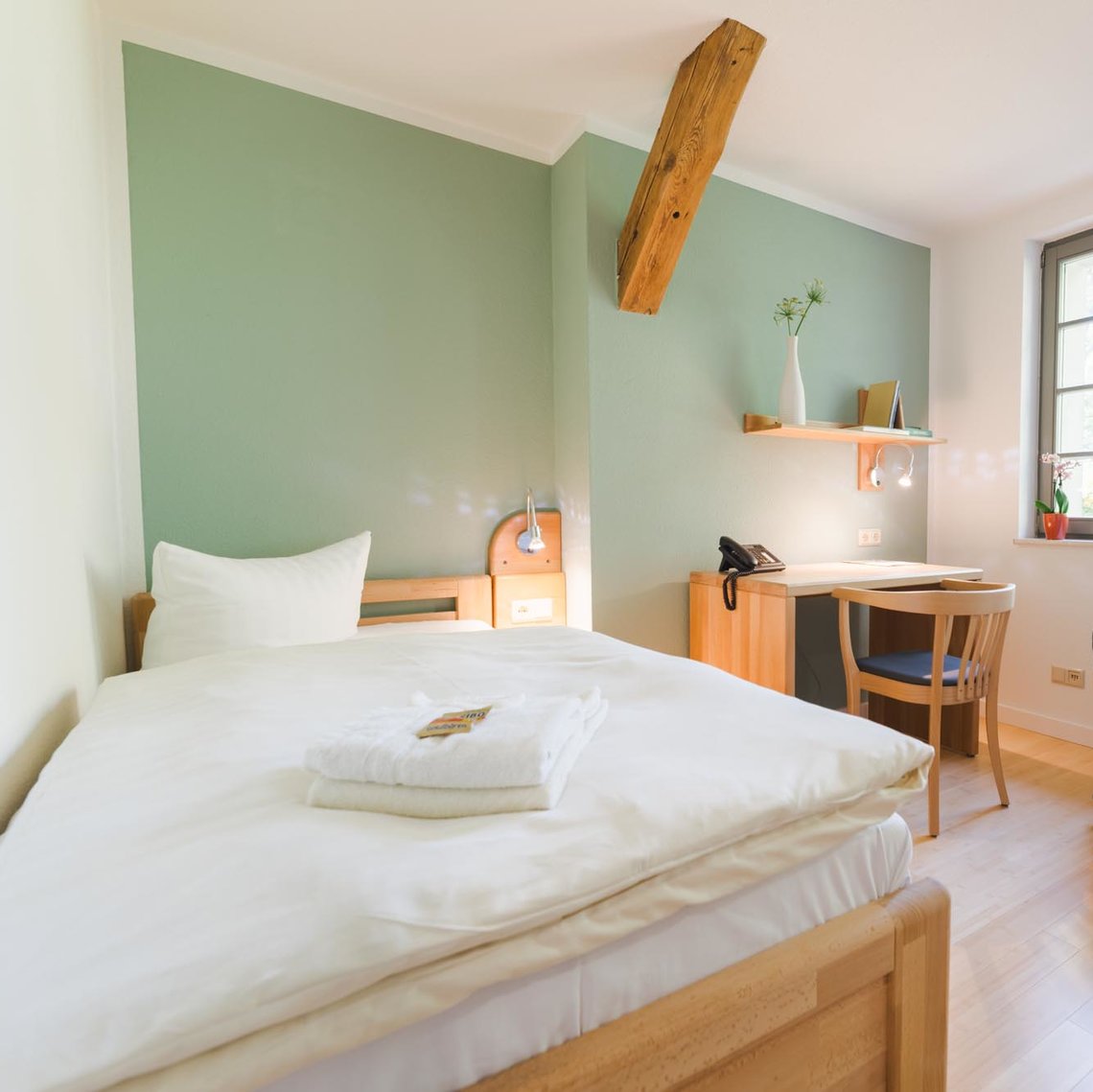 Man sieht ein Hotelzimmer mit links einem Bett mit Handtüchern, in der Mitte einem Schreibtisch mit einem Stuhl und einem Bücherregal darüber und rechts daneben einen grauen Sessel vor zwei Fenstern. Die Mittlere Wand ist grün.