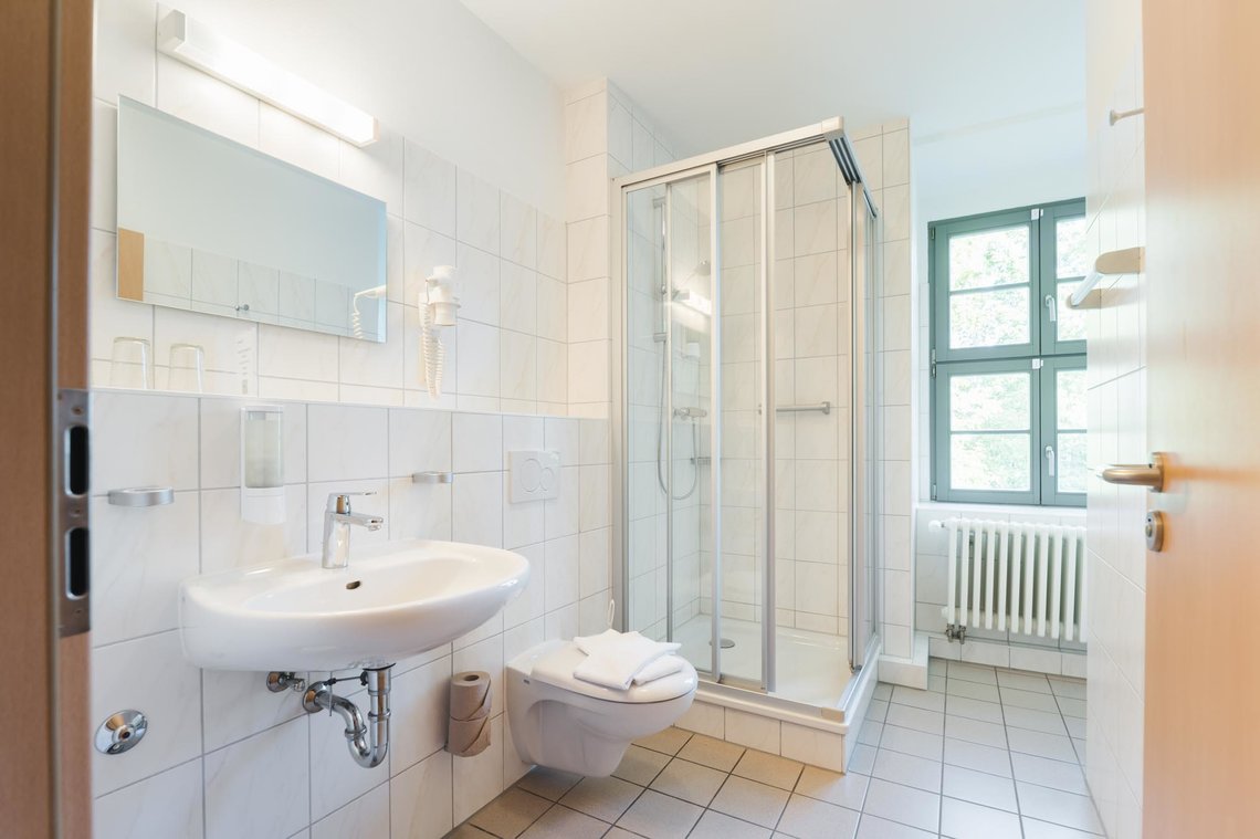 Ein weiß gefliestes Badezimmer mit einem Fenster, einer quadratischen Dusche in der Ecke, einer Toilette mit 2 Handtüchern darauf und einem Waschbecken mit einem Spiegel. Neben dem Spiegel hängt ein Fön in einer Halterung.
