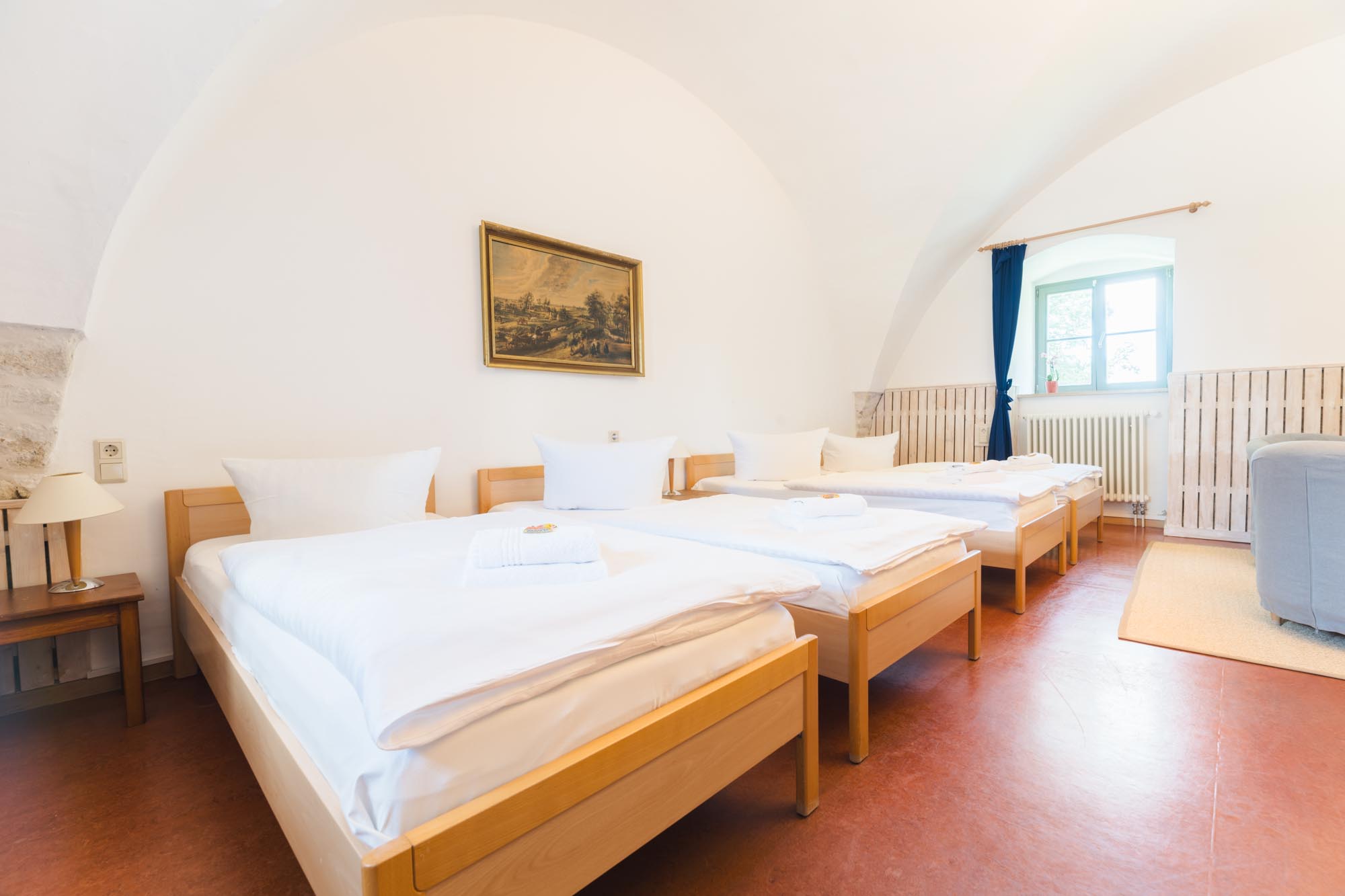 Helles, geräumiges Zimmer mit vier Einzelbetten in einer Reihe. An der Wand hängt ein Gemälde, und ein Fenster mit blauer Gardine lässt Tageslicht herein.