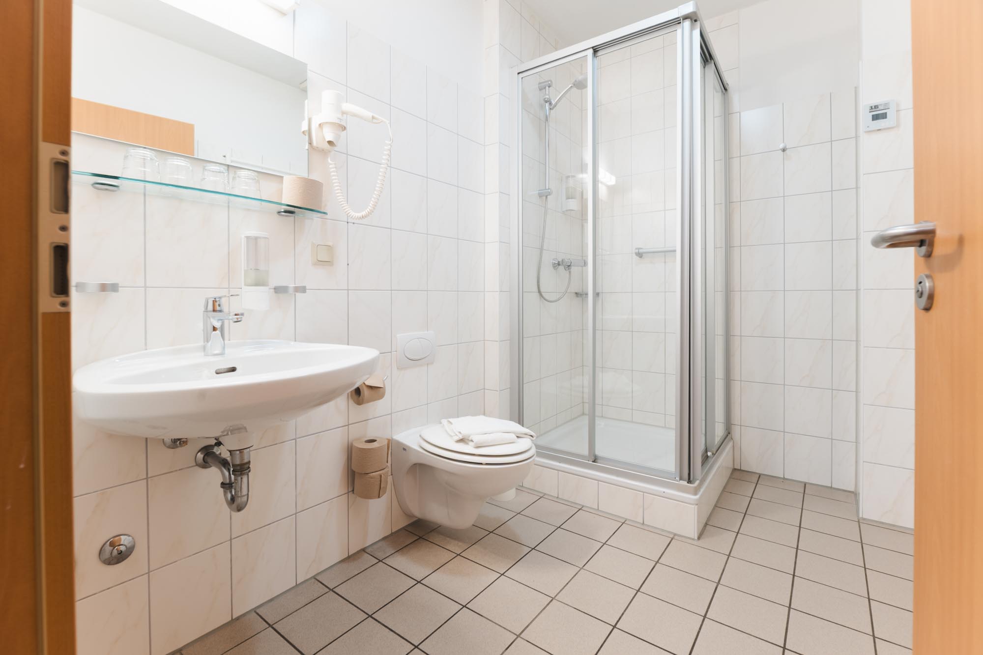 Ein weiß gefliestes Badezimmer mit einer quadratischen Dusche in der Ecke, einer Toilette mit 2 Handtüchern darauf und einem Waschbecken mit einem Spiegel. Neben dem Spiegel hängt ein Fön in einer Halterung.